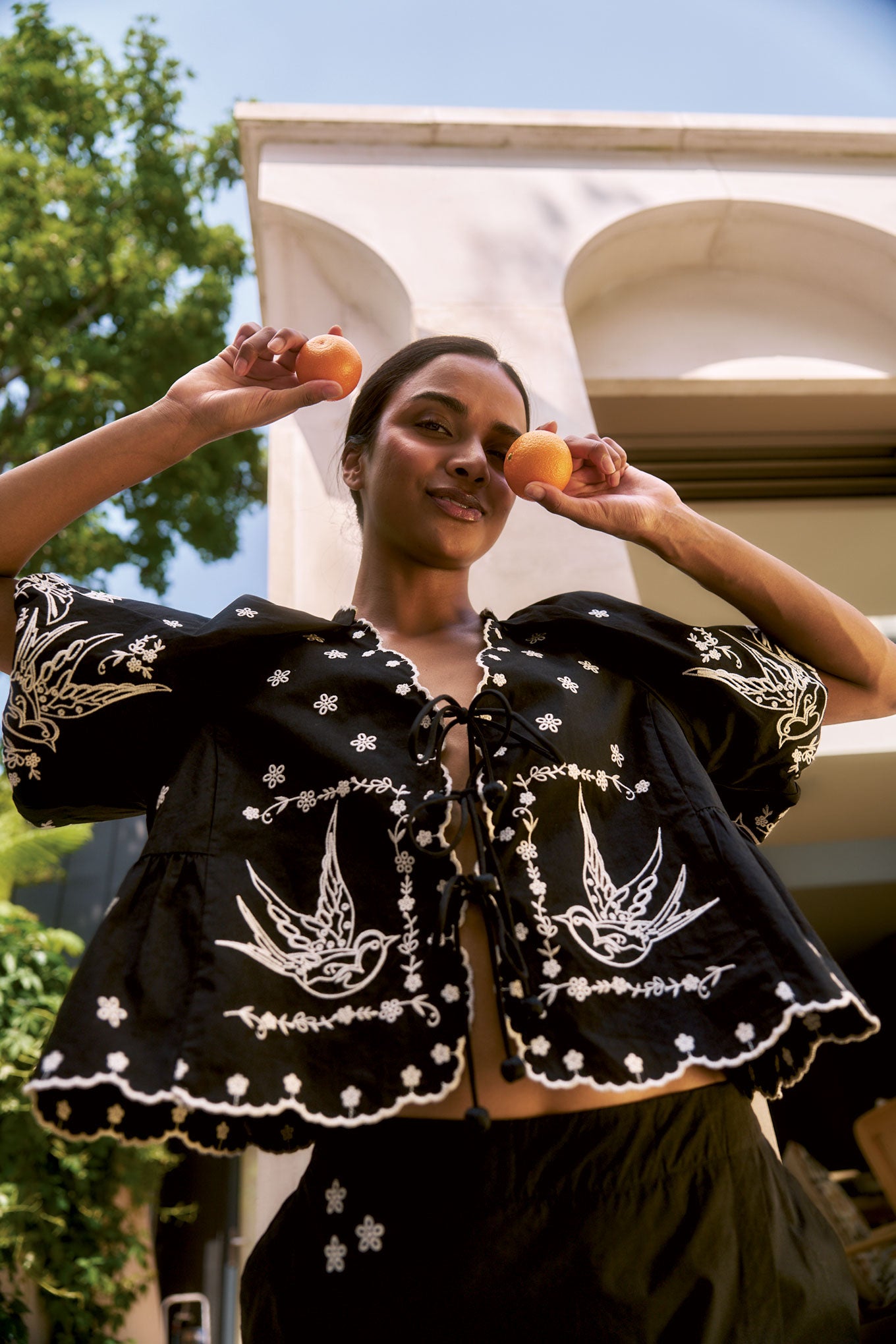 Woman wearing BEATRIX Organic Cotton Top - Black, holding oranges in a sunny outdoor setting.