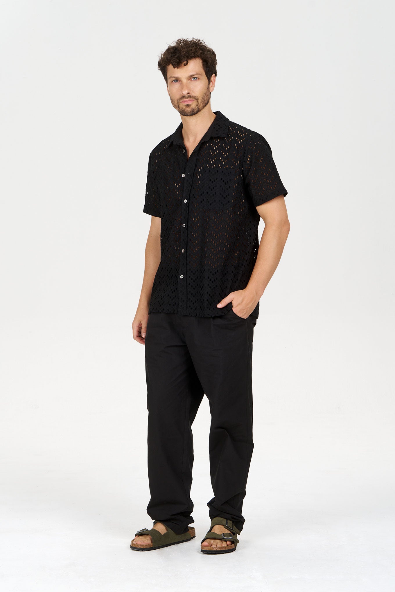 Model wearing BOWIE Organic Cotton Trouser - Black with a black patterned shirt and sandals, showcasing a relaxed look.
