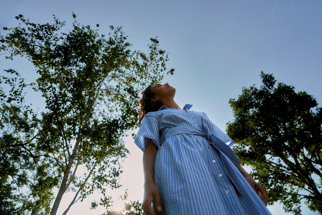 A woman wearing the SHORE Organic Cotton Dress - Blue Stripe standing under tall trees with a clear sky above.