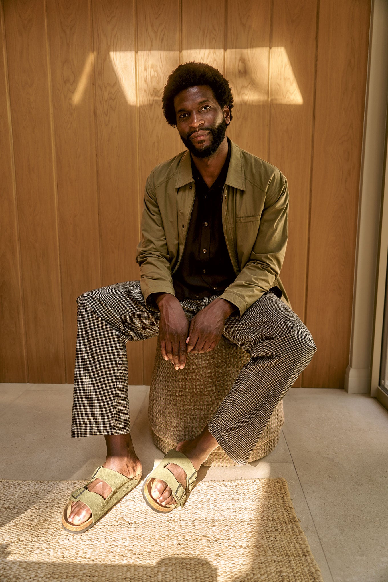 RUDY Organic Cotton Jacket - Olive worn by a man sitting on a stool in a sunlit room, showcasing a casual outfit.